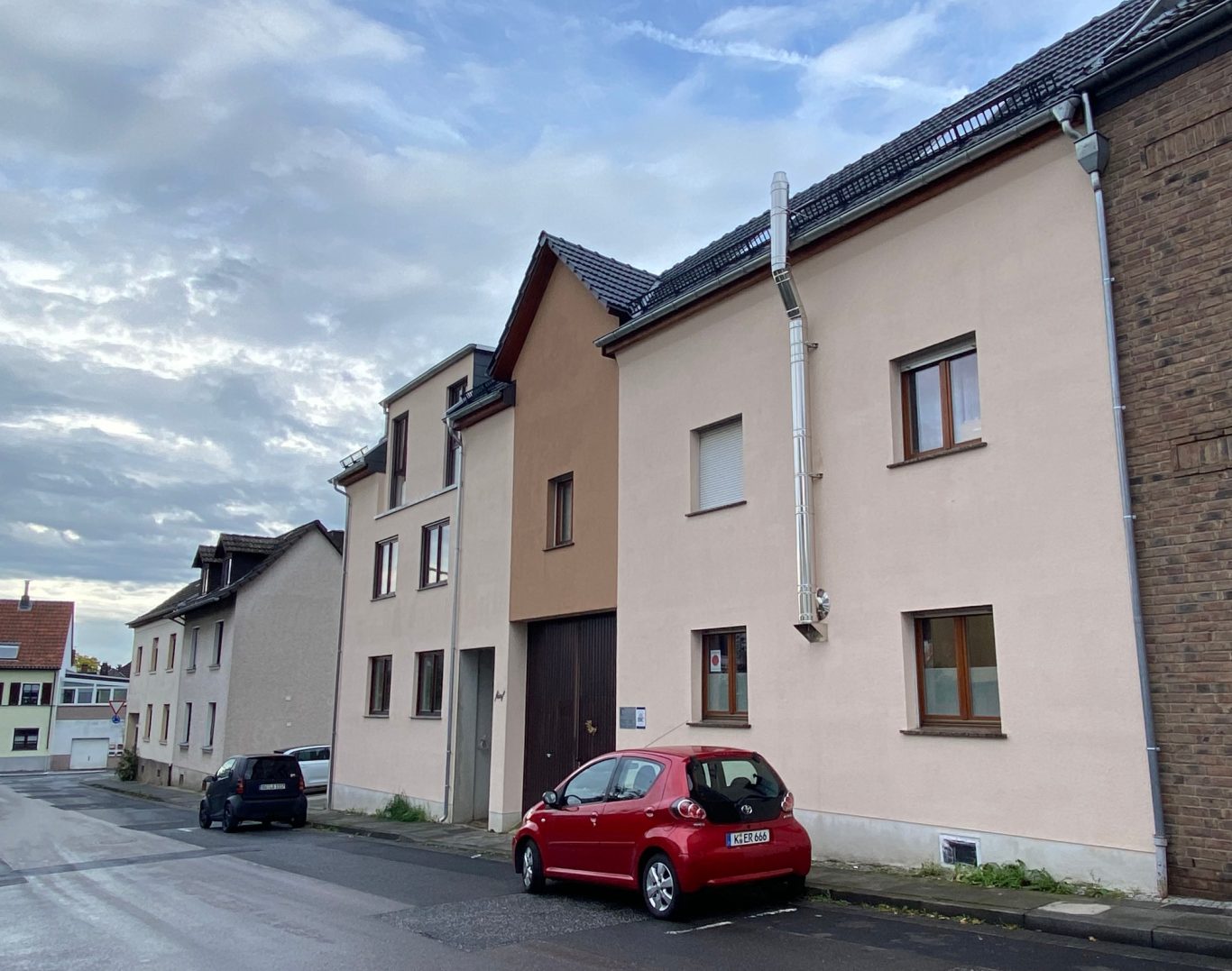 Hausansicht Front Straßenseite Zwei Gebäude an einer Straße mit einem roten Auto davor und bewölktem Himmel.