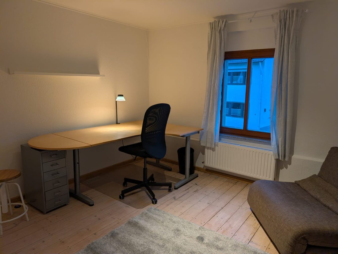Zimmer 2 OG rechts Heller Arbeitsbereich mit Schreibtisch, Stuhl und Fenster, beige Couch im Hintergrund.