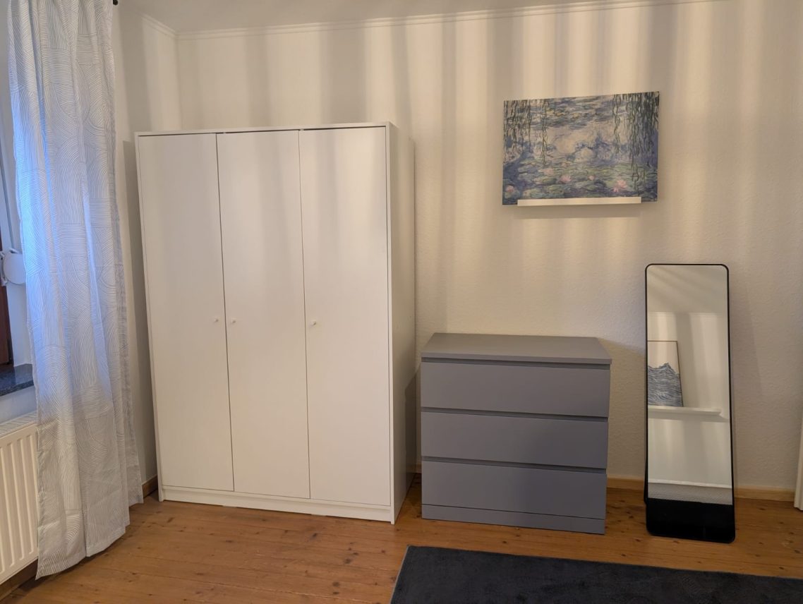 Erdgeschoss links Weißer Kleiderschrank, graue Kommode, Spiegel und ein Bild an der Wand.