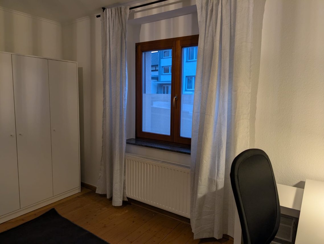 Erdgeschoss rechts Ein heller Raum mit Fenster, Vorhängen, Schreibtisch und schwarzem Stuhl.