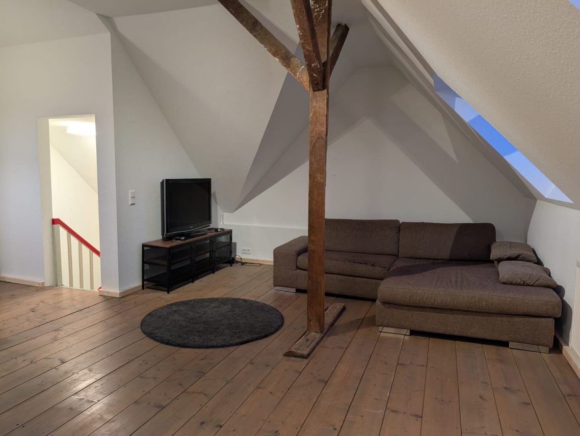 Dachgeschoss Aufenthaltsbereich links Geräumiger Dachboden mit Holzfußboden, Sofa, Fernseher und Treppe.