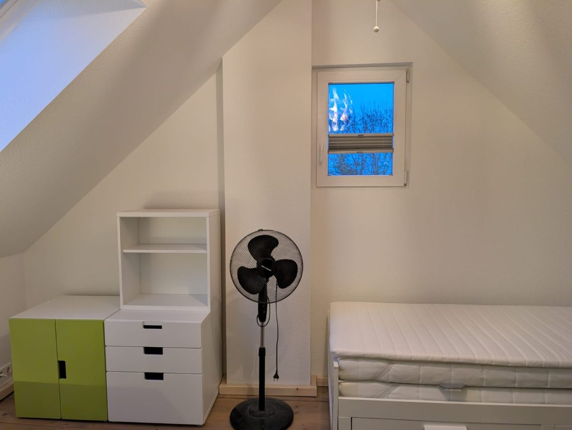 Zimmer Dachgeschoss rechts Schlichtes Zimmer mit Bett, weißem Schrank und Ventilator, Fenster mit blauer Sicht.