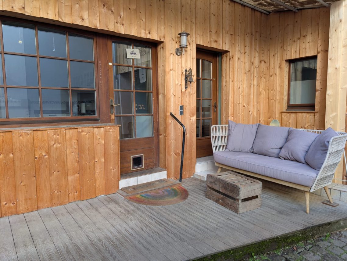 Eingang zur WG Holzhaus mit zwei Fenstern, Eingangstüren und einer Bank auf der Veranda.