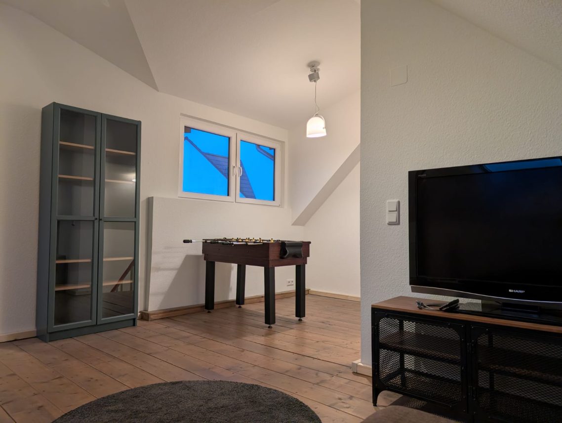 Dachgeschoss Aufenthaltsbereich rechts Heller Raum mit Fenstern, einem Tisch, Regalen und einem Fernseher.