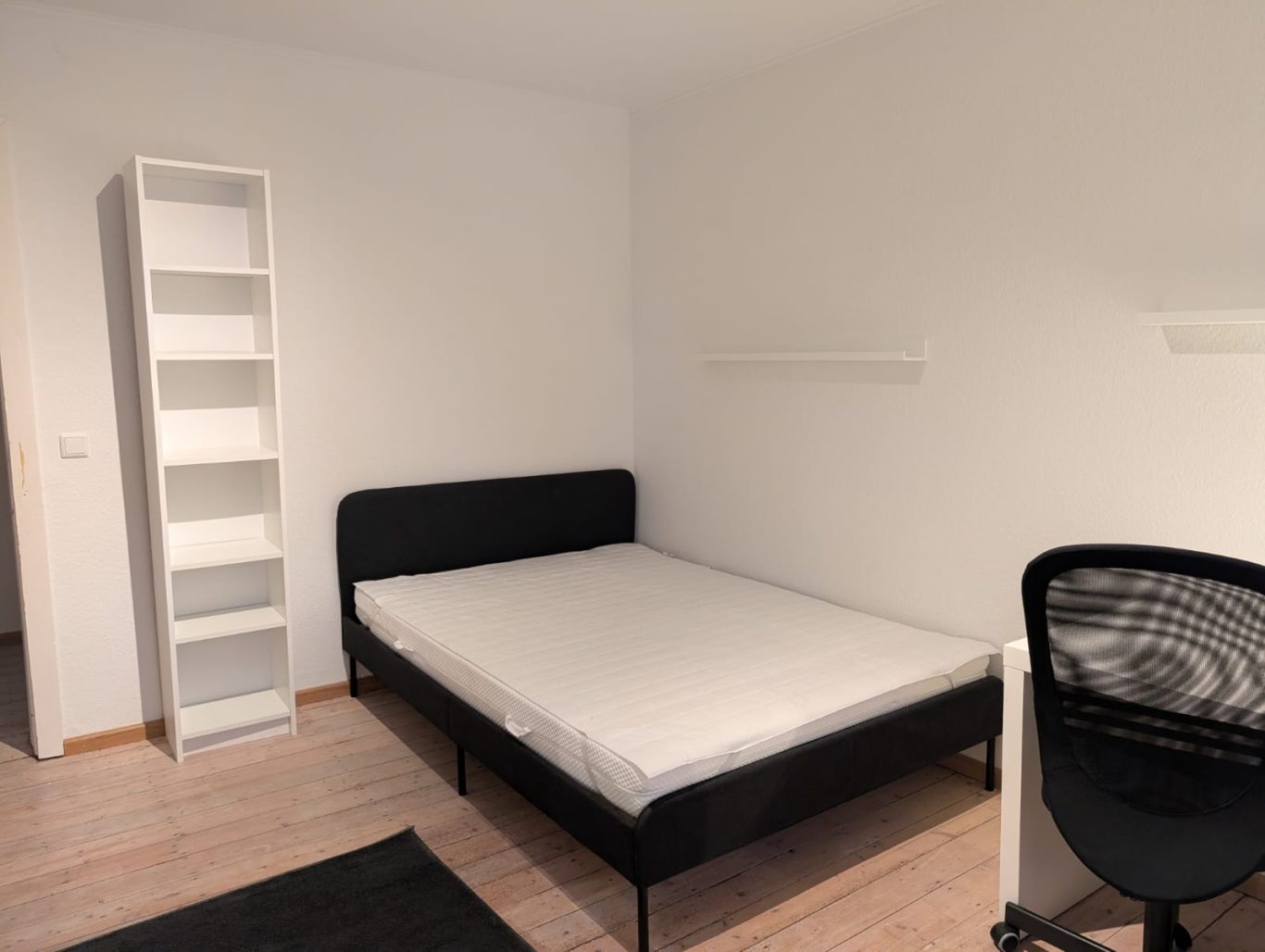 Zimmer OG links Schlichtes Schlafzimmer mit Bett, Regal und Bürostuhl auf hellem Boden.